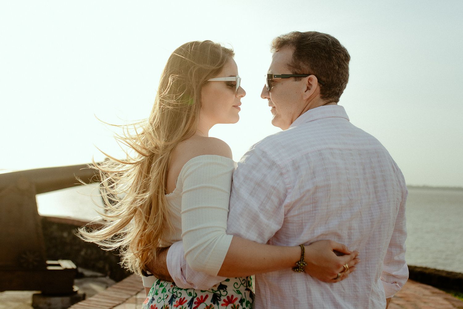 A romantic couple embraces on a beach at sunset with the woman's hair blowing in the wind.