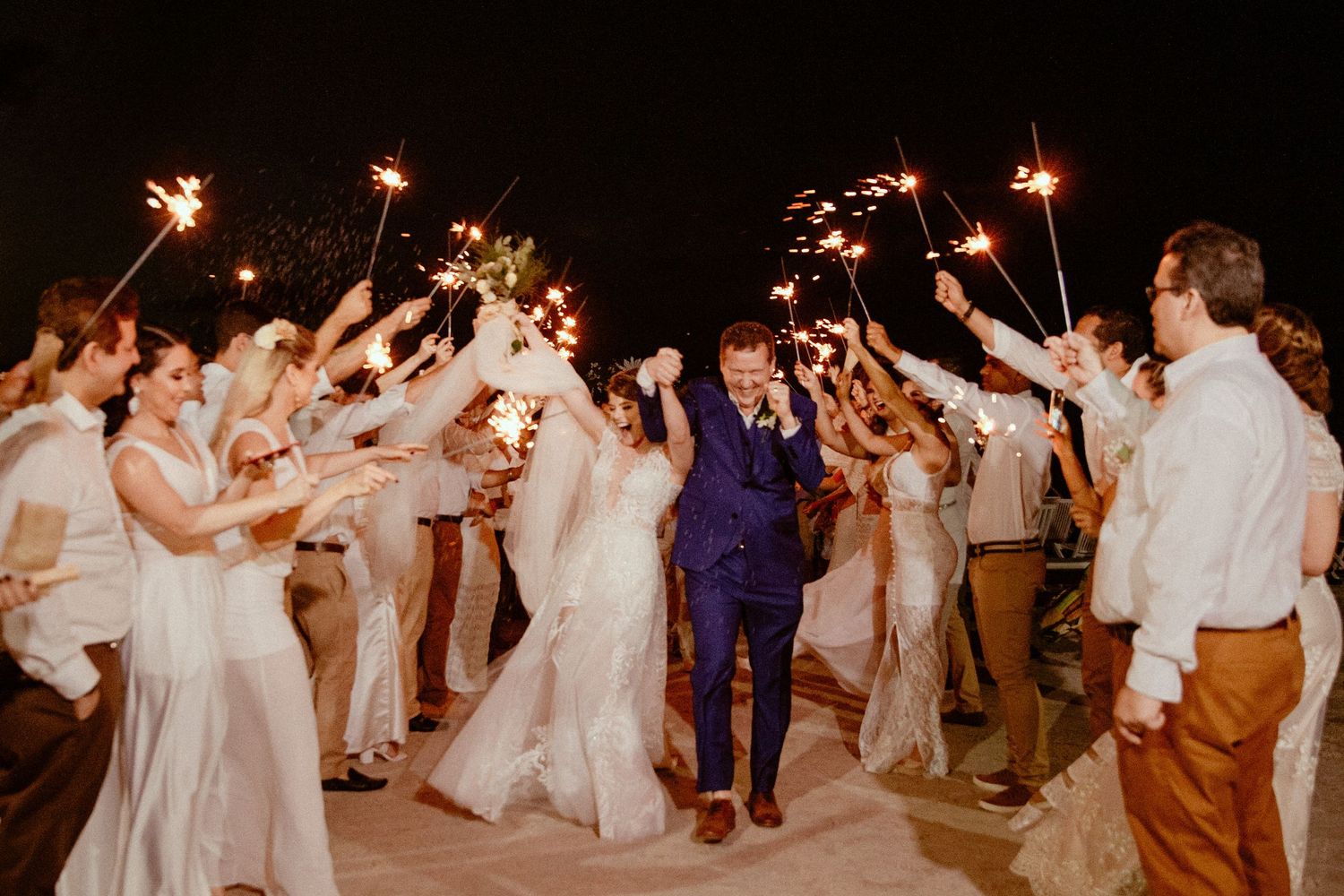 Wedding guests dance and celebrate with sparklers held high during an energetic nighttime reception celebration.