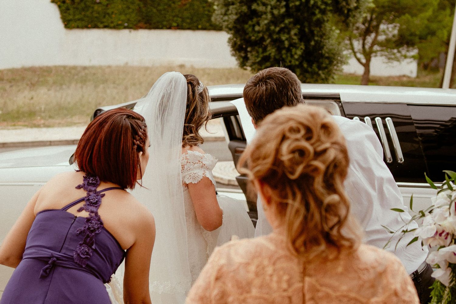 Group of bridesmaids in plum-colored dresses help adjust a white wedding gown during outdoor pre-ceremony preparations.