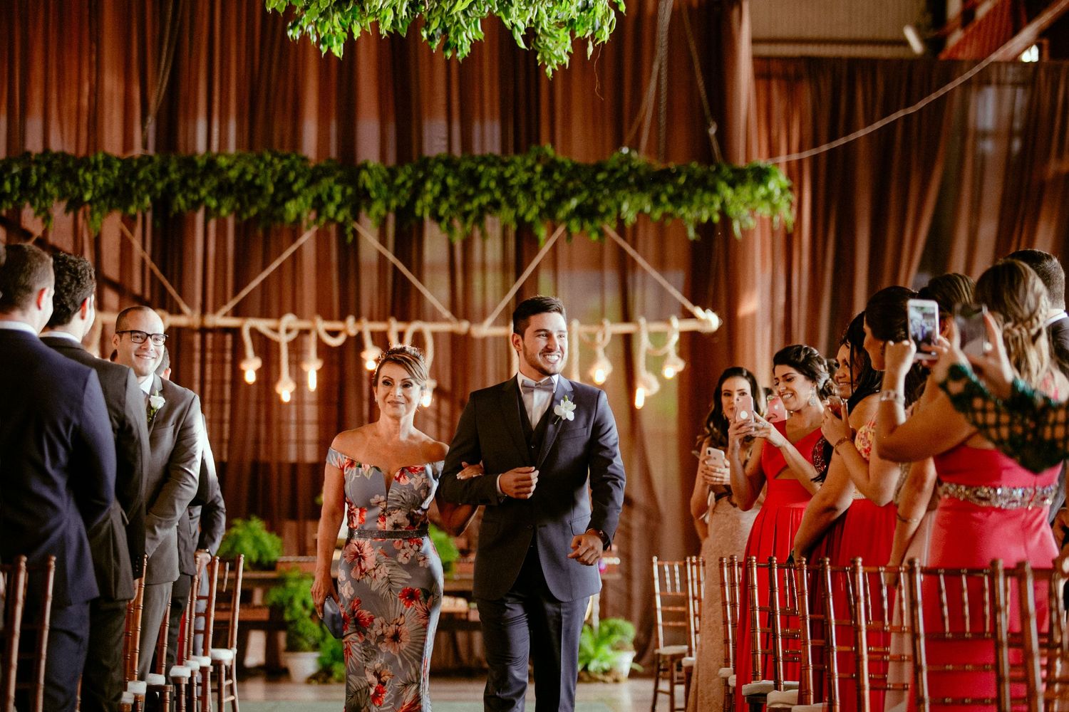 Wedding guests stand in rows during an indoor ceremony under greenery at a rustic wooden venue.