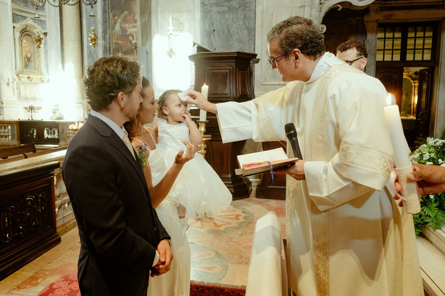 Priest performing a baptism ceremony on an infant held by parents inside an ornate Catholic church.