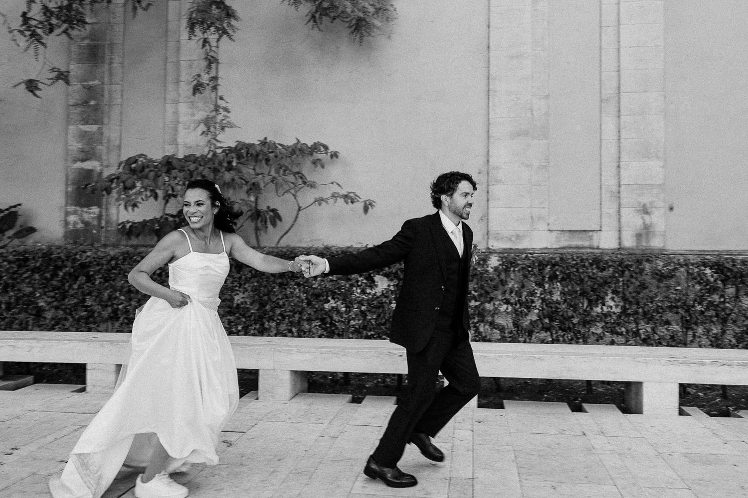 Black and white photo of a bride and groom joyfully running hand in hand outdoors on their wedding day.