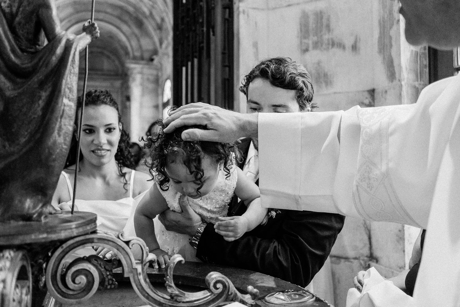 Black and white photo of a Catholic baptism ceremony with a priest placing hand on a curly-haired child's head.