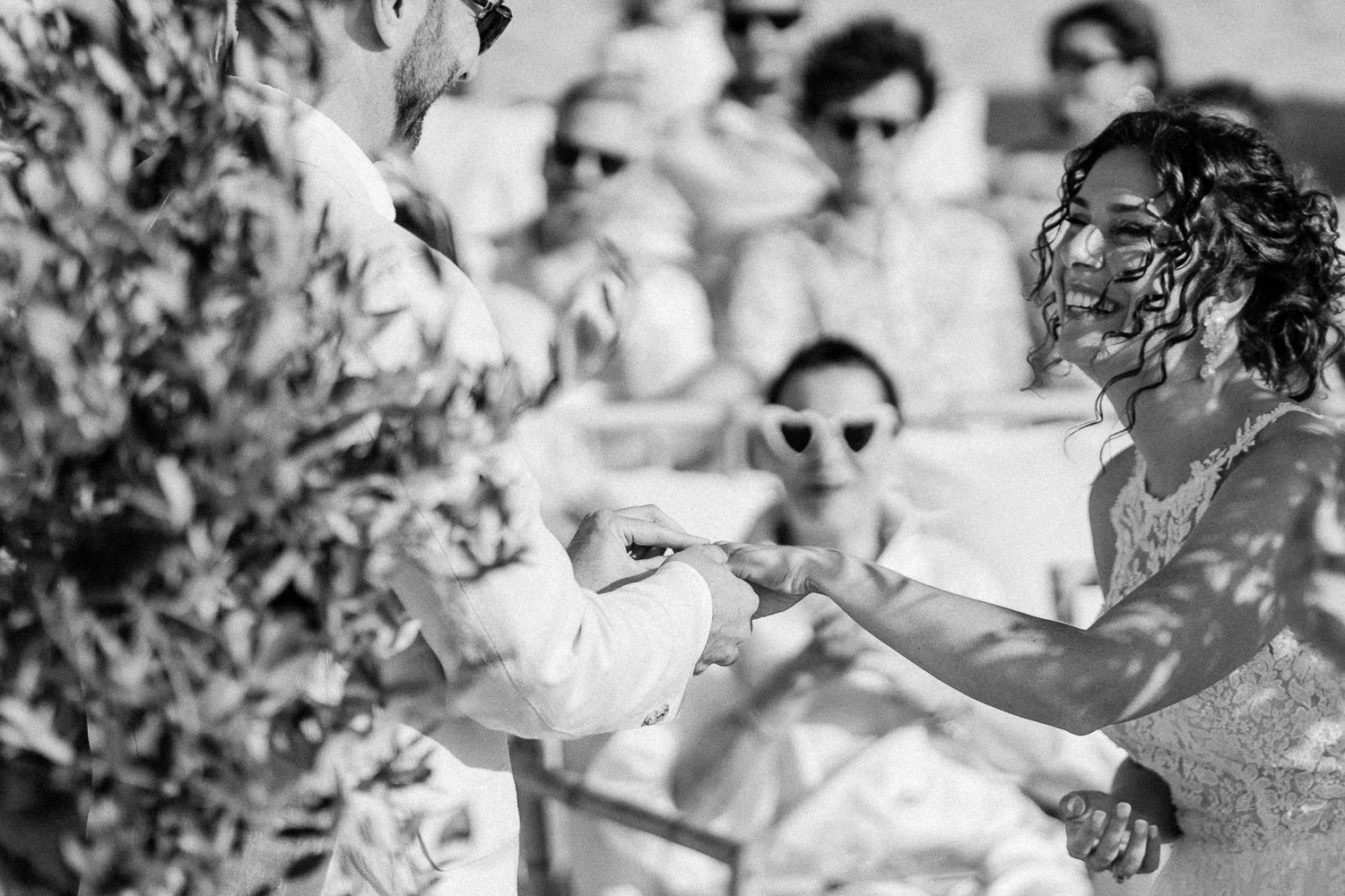 Black and white wedding photo of bride exchanging rings outdoors with guests wearing sunglasses in background.