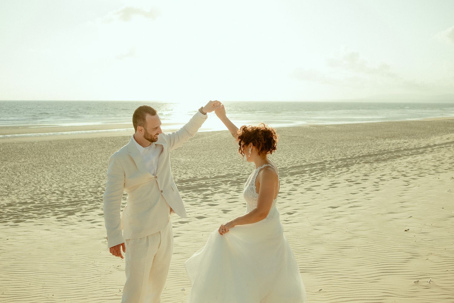 Bride and groom dancing on a sandy beach, wearing white wedding attire with ocean waves in the background.