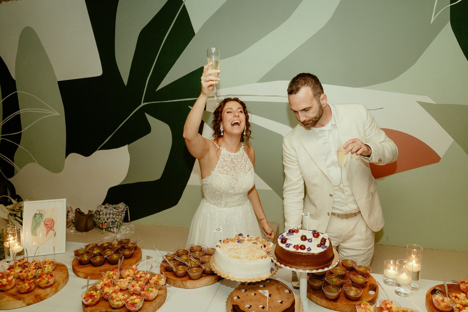 Bride and groom laughing while cutting wedding cake at dessert table with candles and tropical wall art.