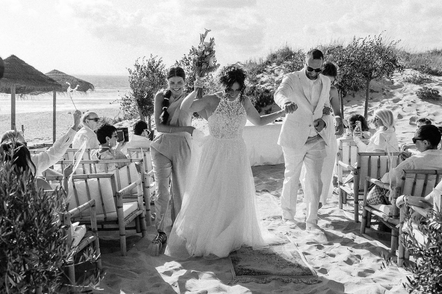 Black and white beach wedding ceremony with bride in flowing gown assisted by guests on sandy shore.