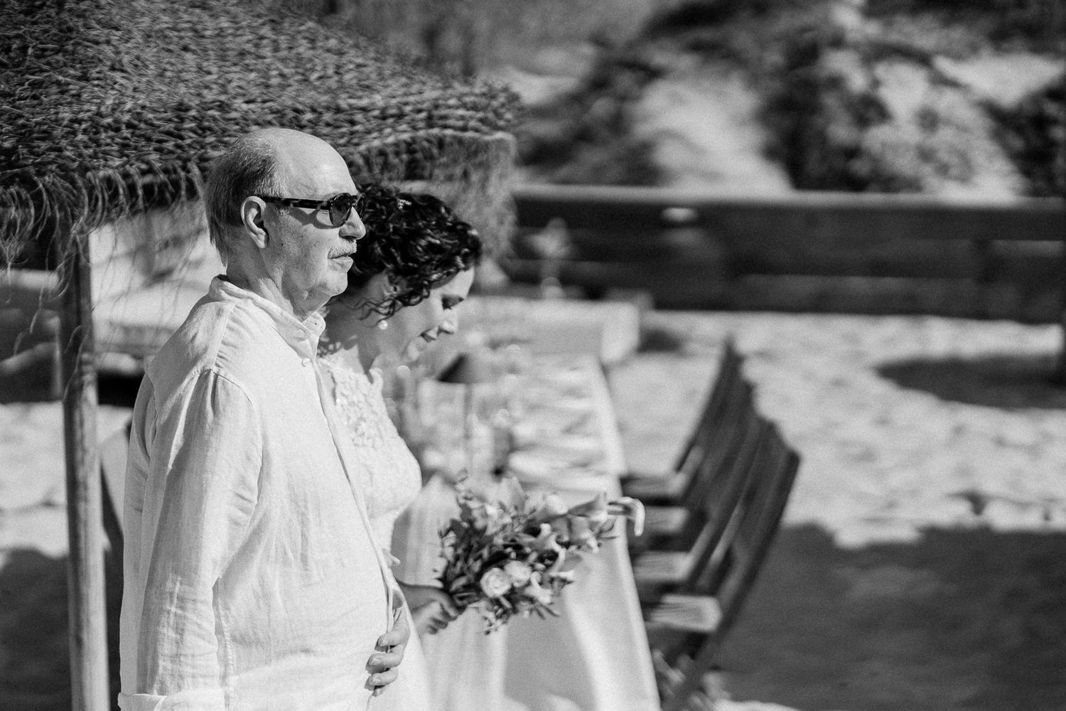 Black and white photo of a bride holding a bouquet walking with an older man at a beach wedding ceremony.