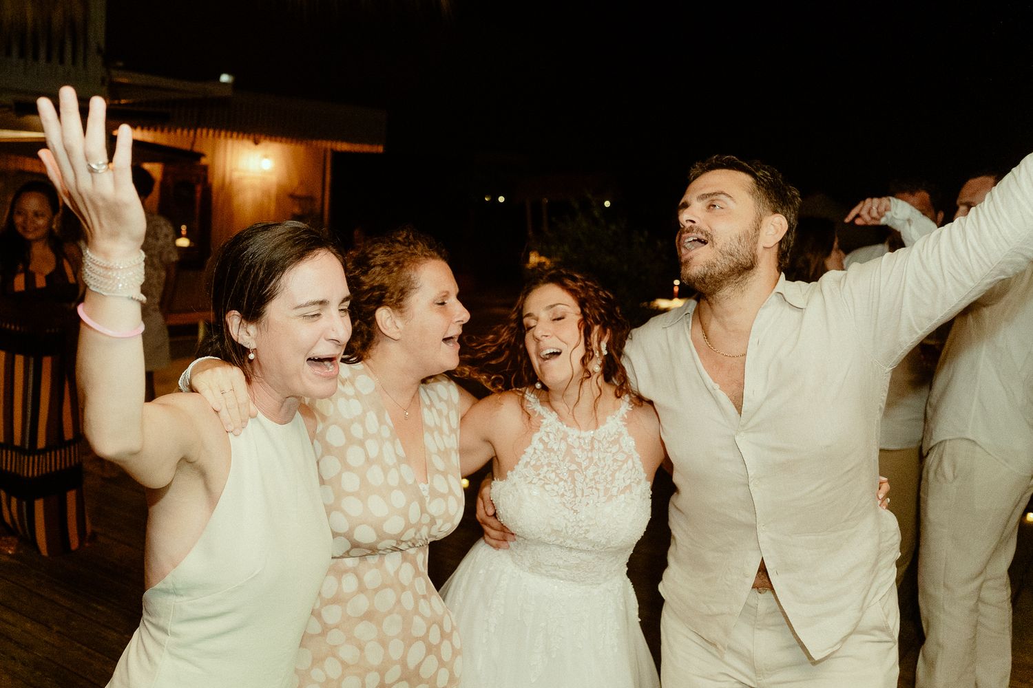 Four friends in white outfits laughing and dancing together at an outdoor evening wedding reception celebration.
