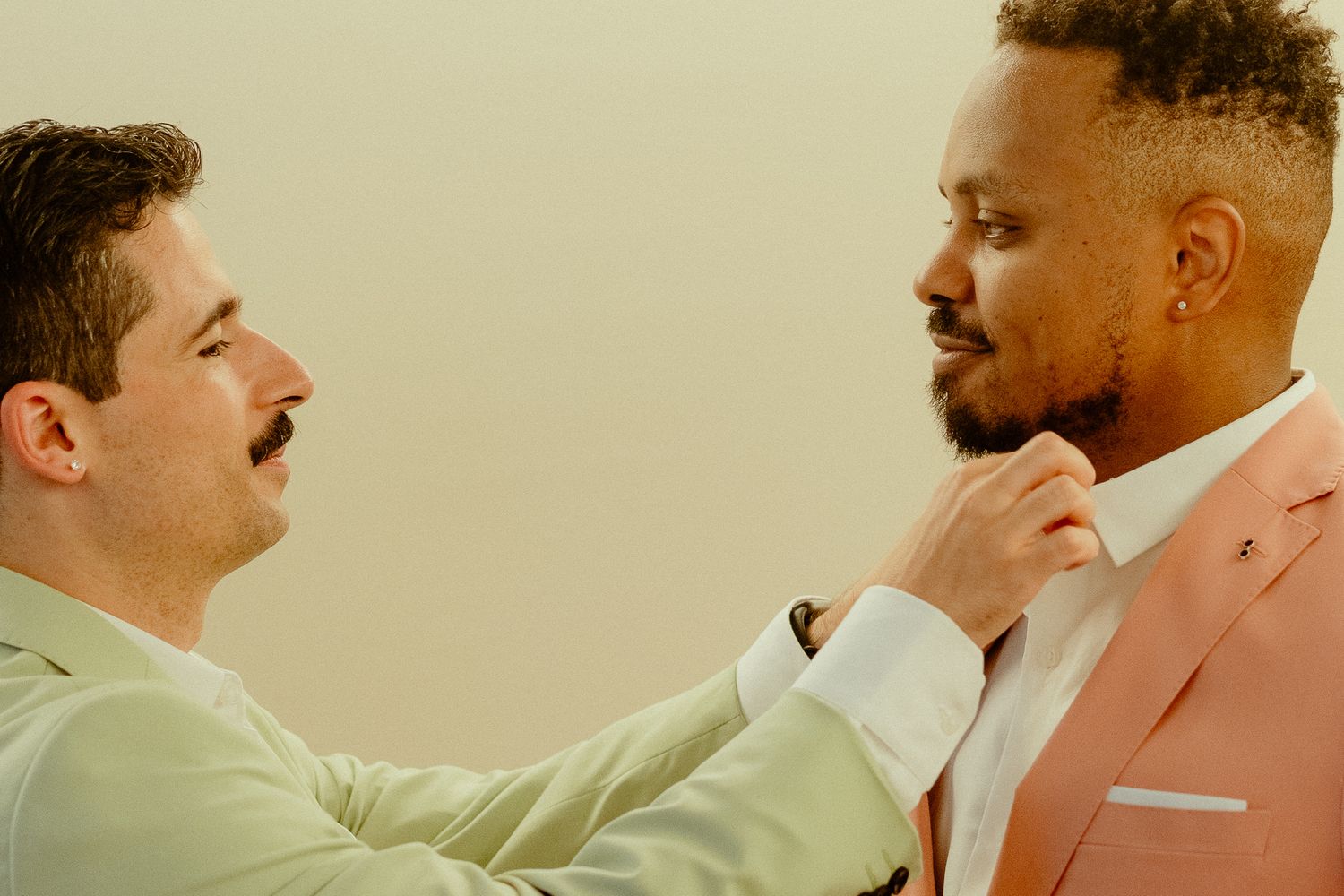 Two men in pastel suits adjusting a tie, sharing a tender moment against a neutral background.