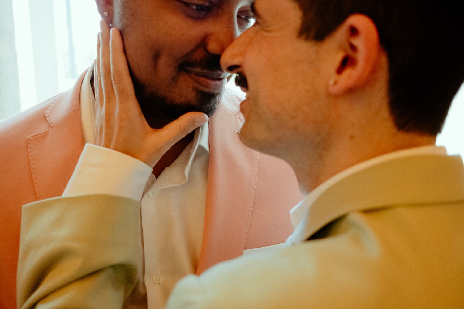 Two men in suits share an intimate moment, tenderly touching each other's faces in a romantic wedding setting.