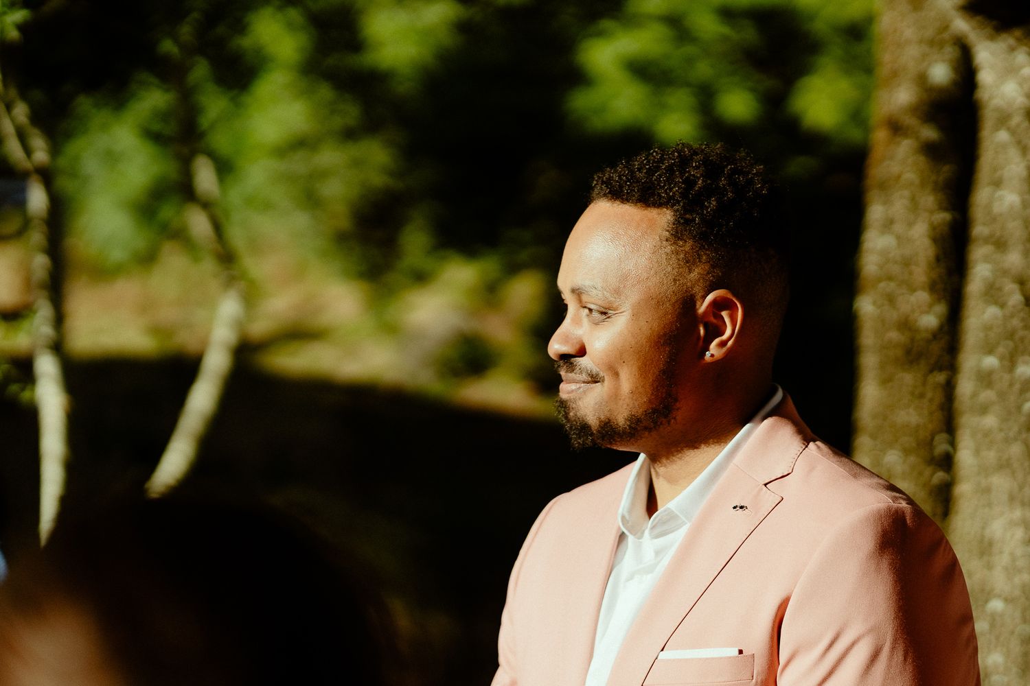 Man in pink blazer standing outdoors among trees, looking thoughtfully to the side in natural light.