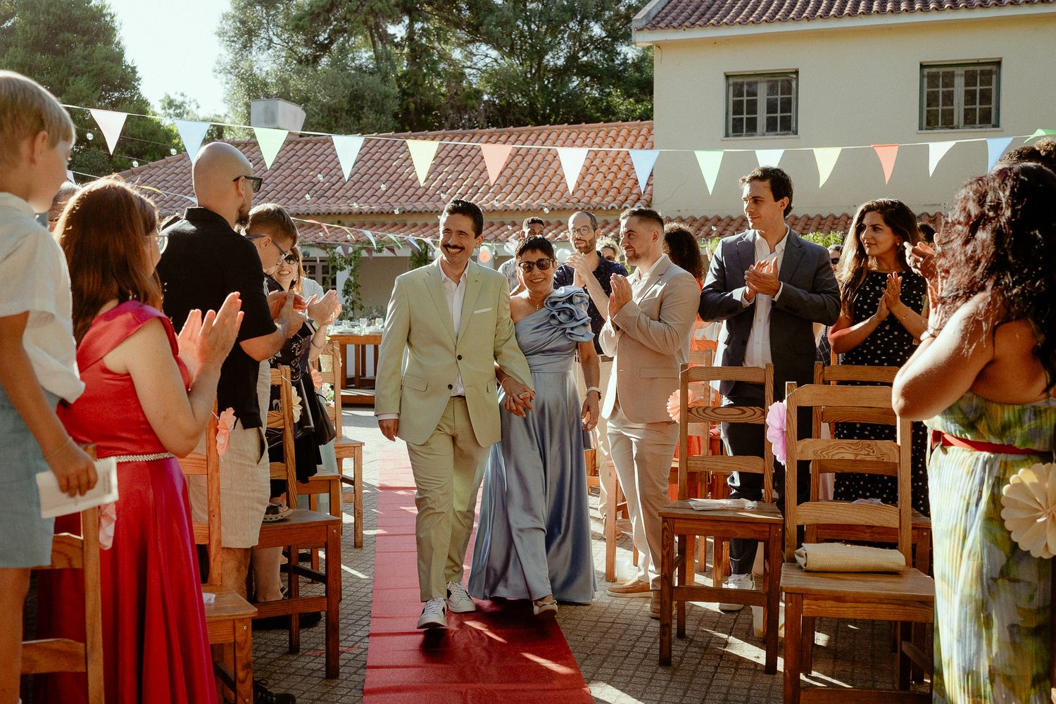 Outdoor wedding ceremony with guests applauding as couple walks down aisle, colorful bunting and rustic venue in background.