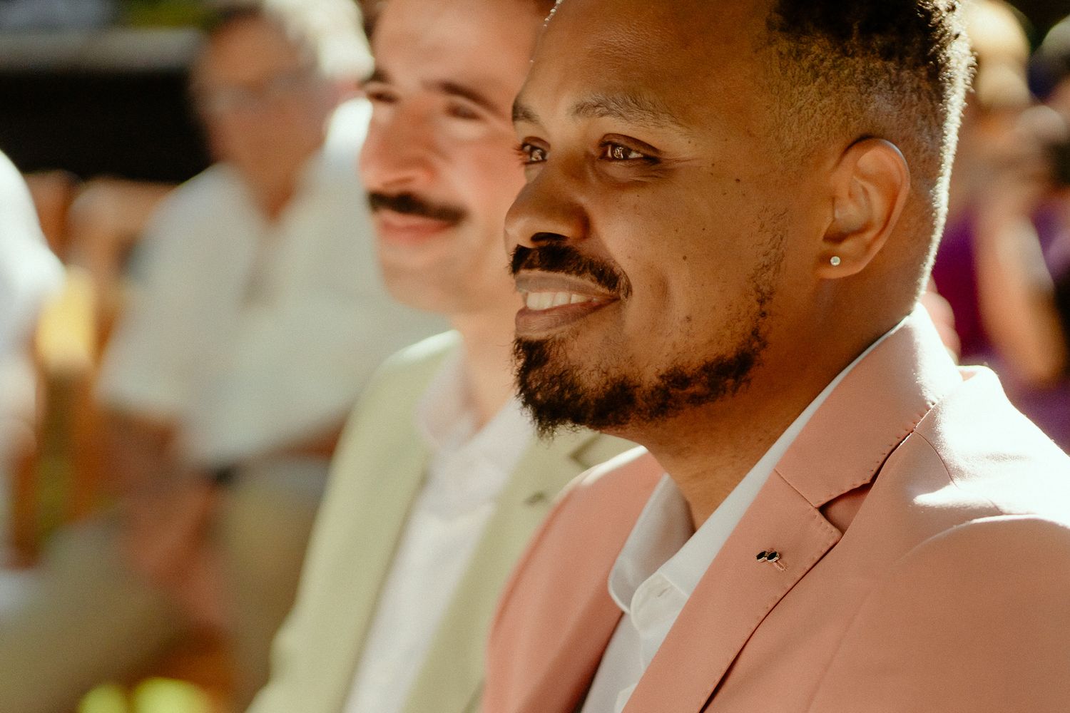 Smiling man in a pink blazer at an outdoor event, with guests visible in the background.