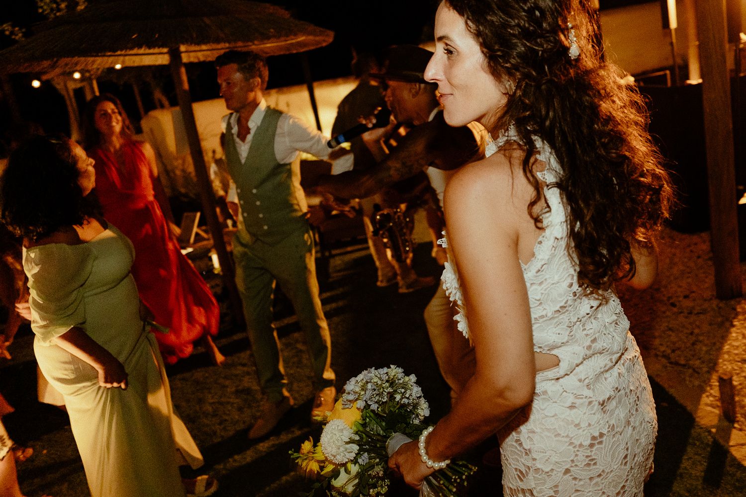 Bride in white lace dress holding bouquet at outdoor wedding reception with guests dancing at night.