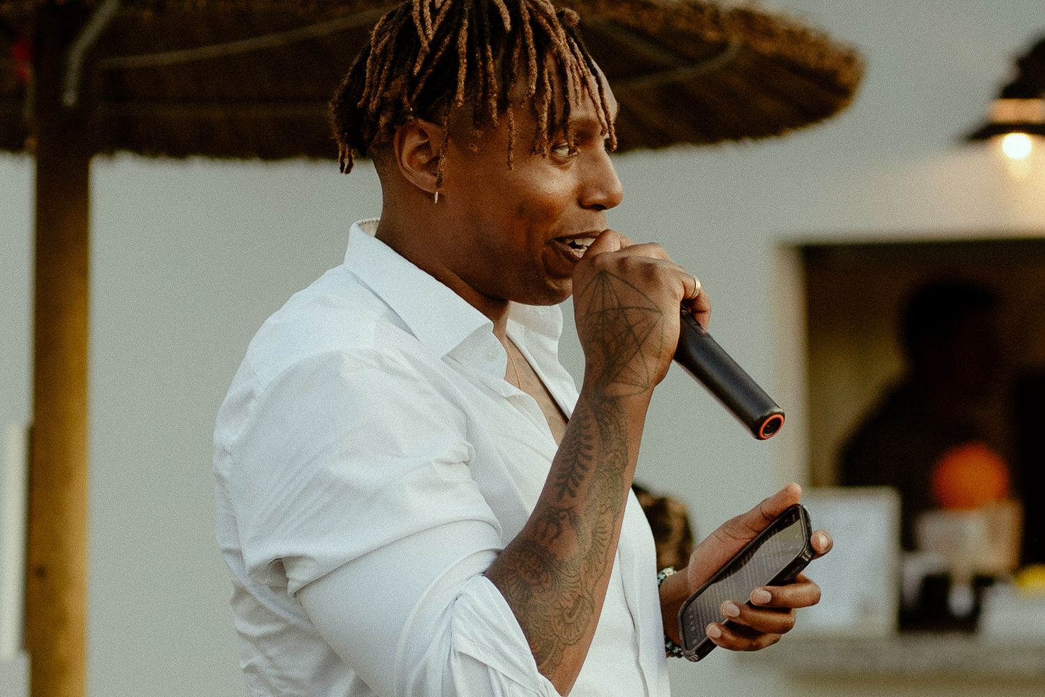 Young man with dreadlocks singing into a microphone outdoors, wearing a white shirt at an event.