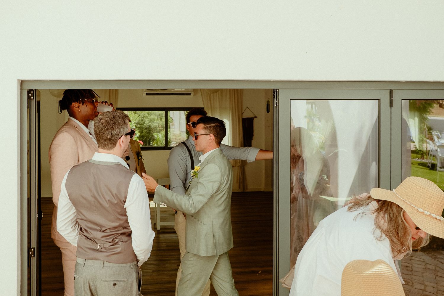 Groomsmen in light suits gather near open glass doors at an outdoor wedding venue on a sunny day.
