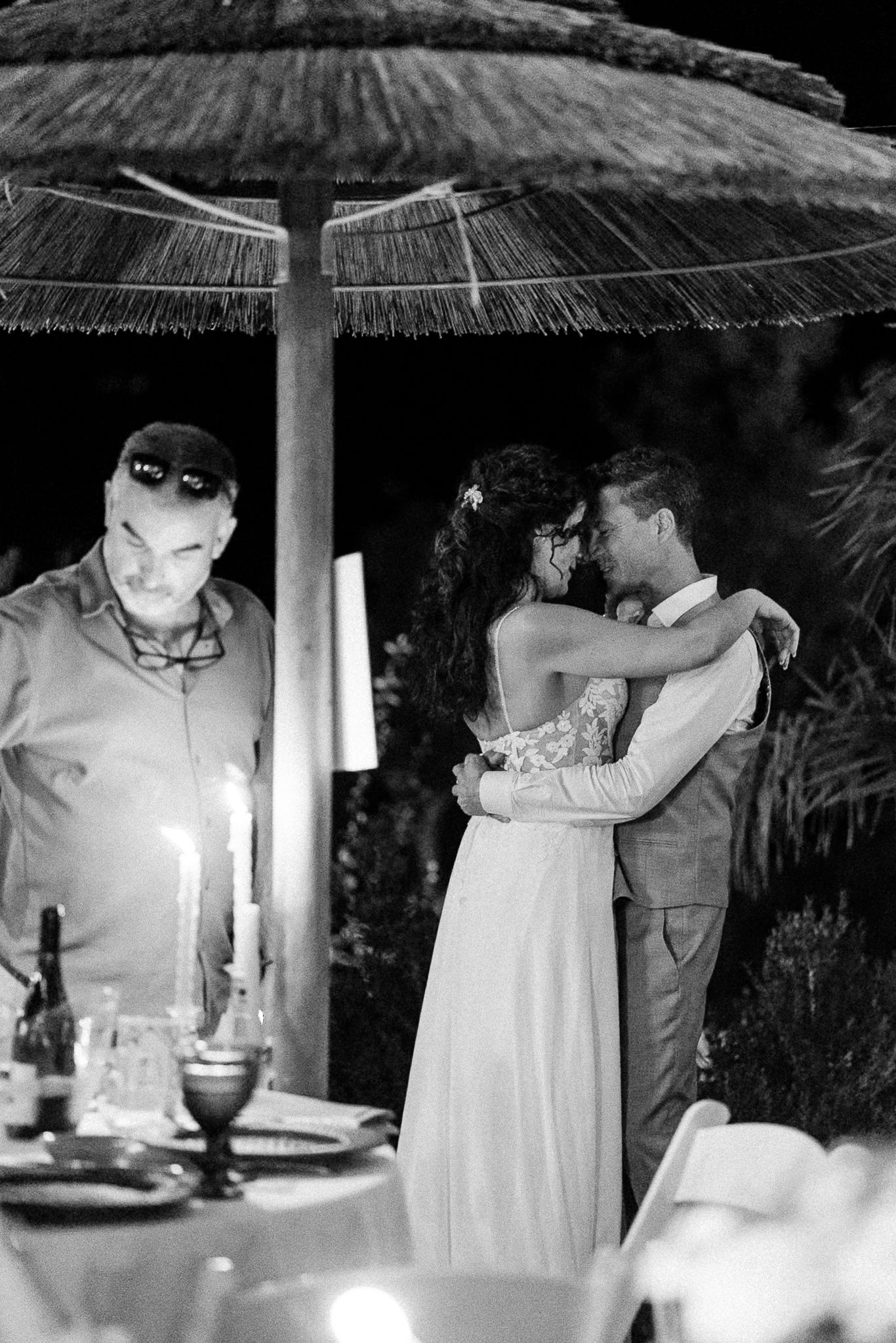 Black and white photo of a couple sharing a romantic embrace at an outdoor candlelit wedding reception at night.