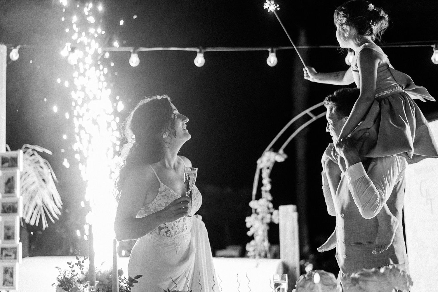 Black and white wedding photo of bride and groom celebrating with sparklers and a child at an outdoor evening reception.