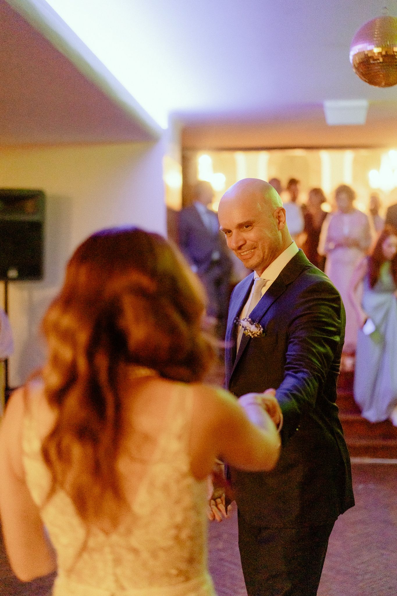 Groom in dark suit smiling at bride during first dance at wedding reception with guests watching.