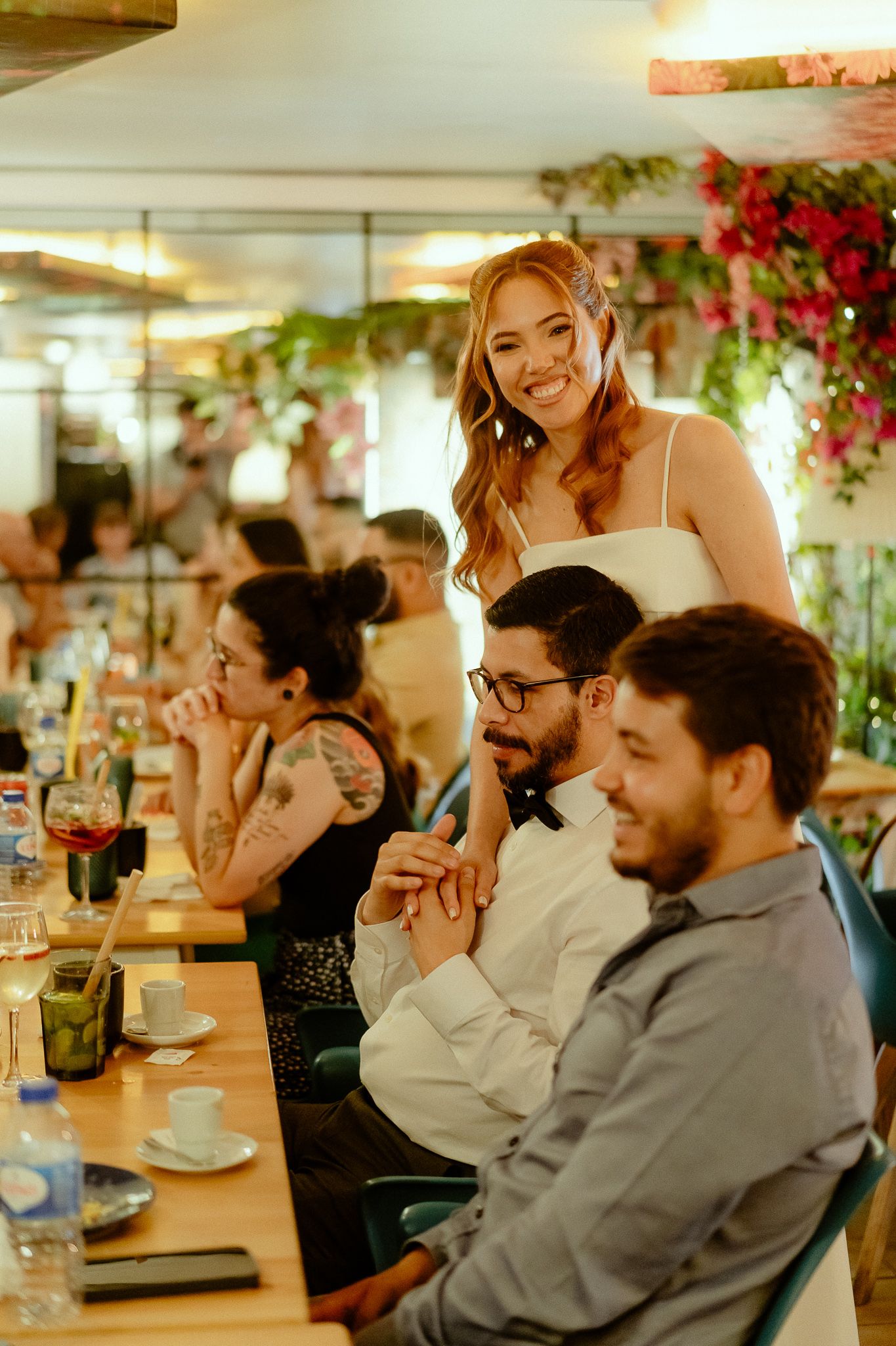 Smiling bride stands behind seated wedding guests at a decorated reception venue with floral arrangements.