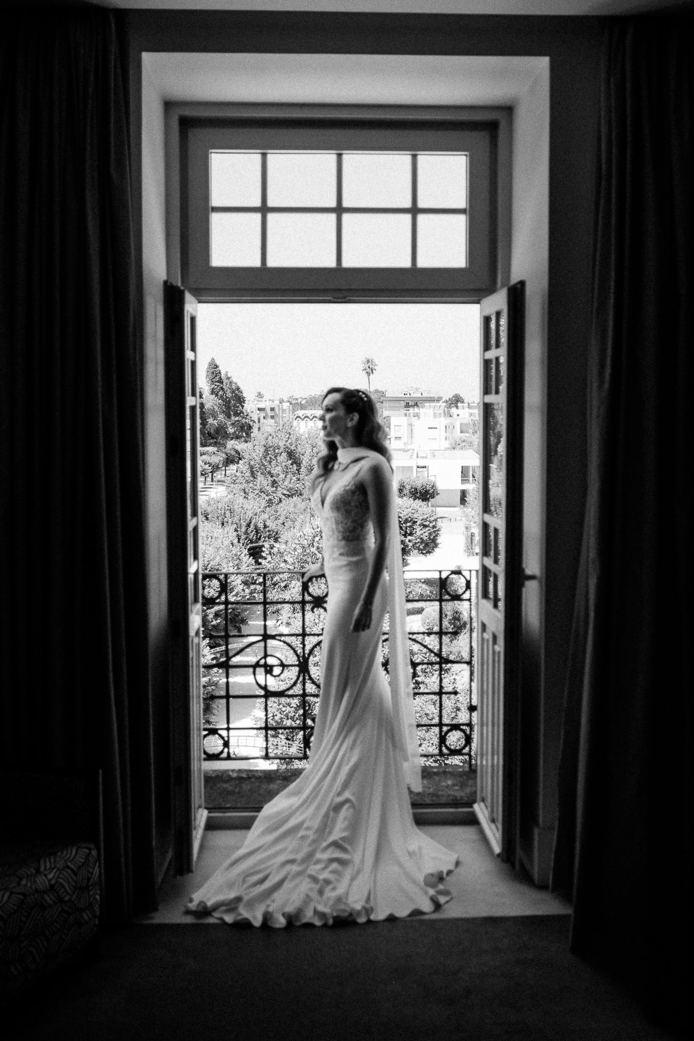 Bride in elegant white gown standing in open doorway with wrought iron balcony and garden view, black and white photo.