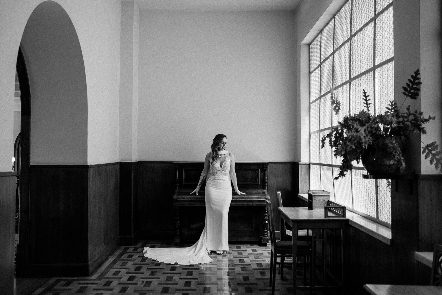 Bride in elegant white gown stands by large window in black and white photo, showcasing timeless wedding dress style.