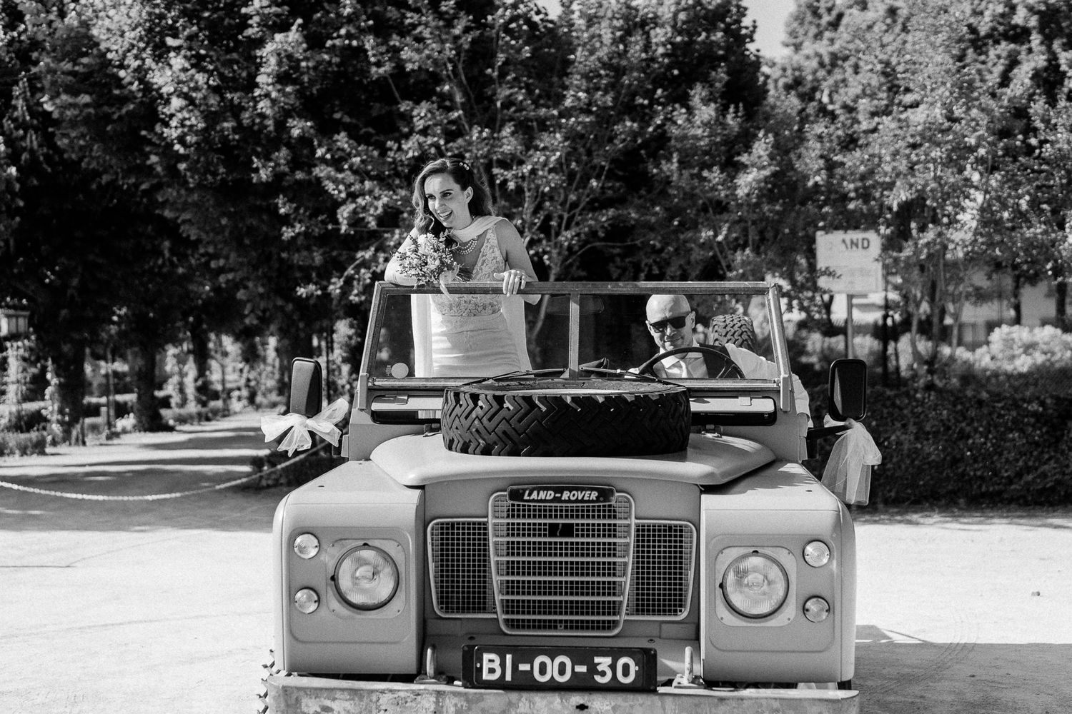 Black and white photo of a couple riding in a vintage Land Rover convertible on a sunny tree-lined road.