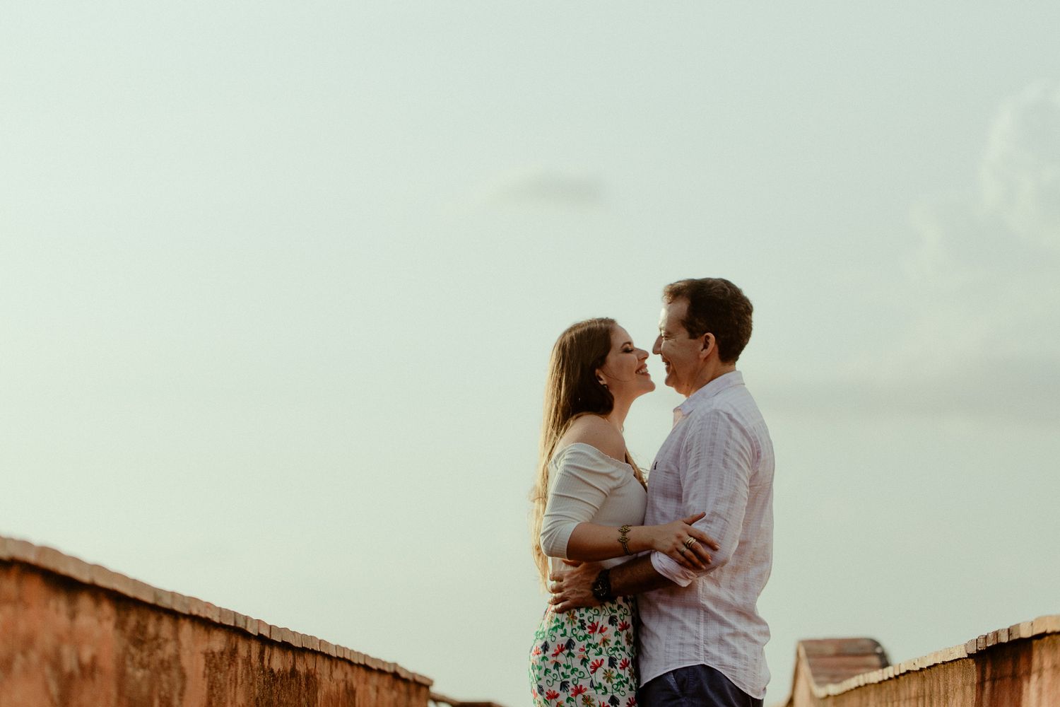 A romantic couple embraces on a rooftop during golden hour against a soft pastel sky.