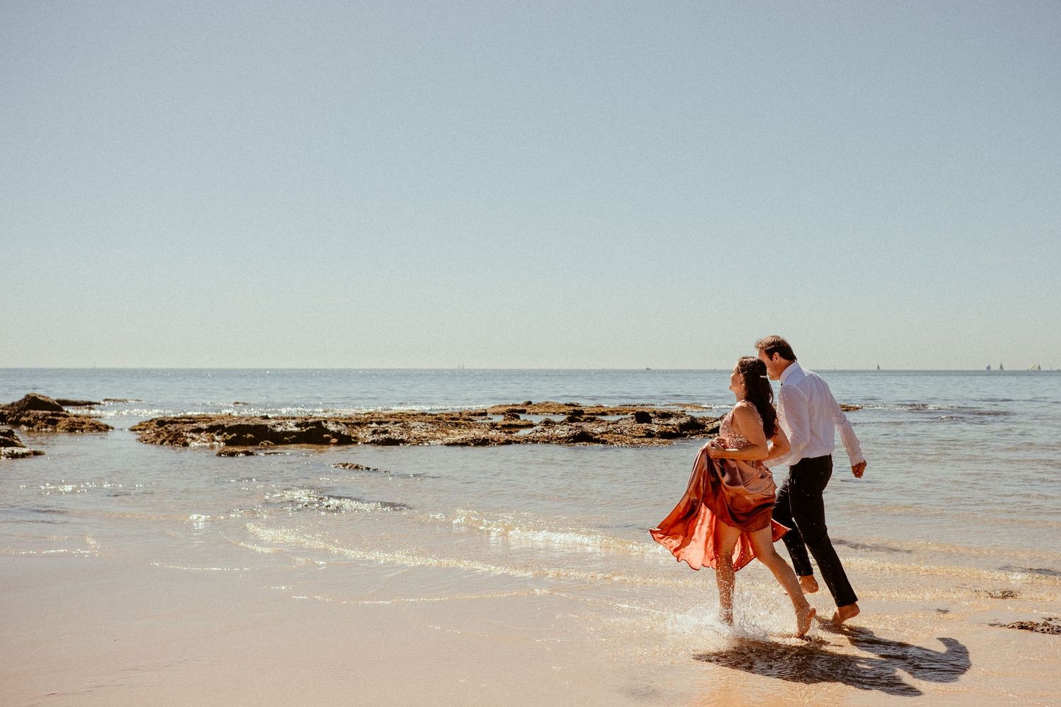 Two people playfully splashing and walking along a sunny beach shoreline with rocky outcrops in the background.