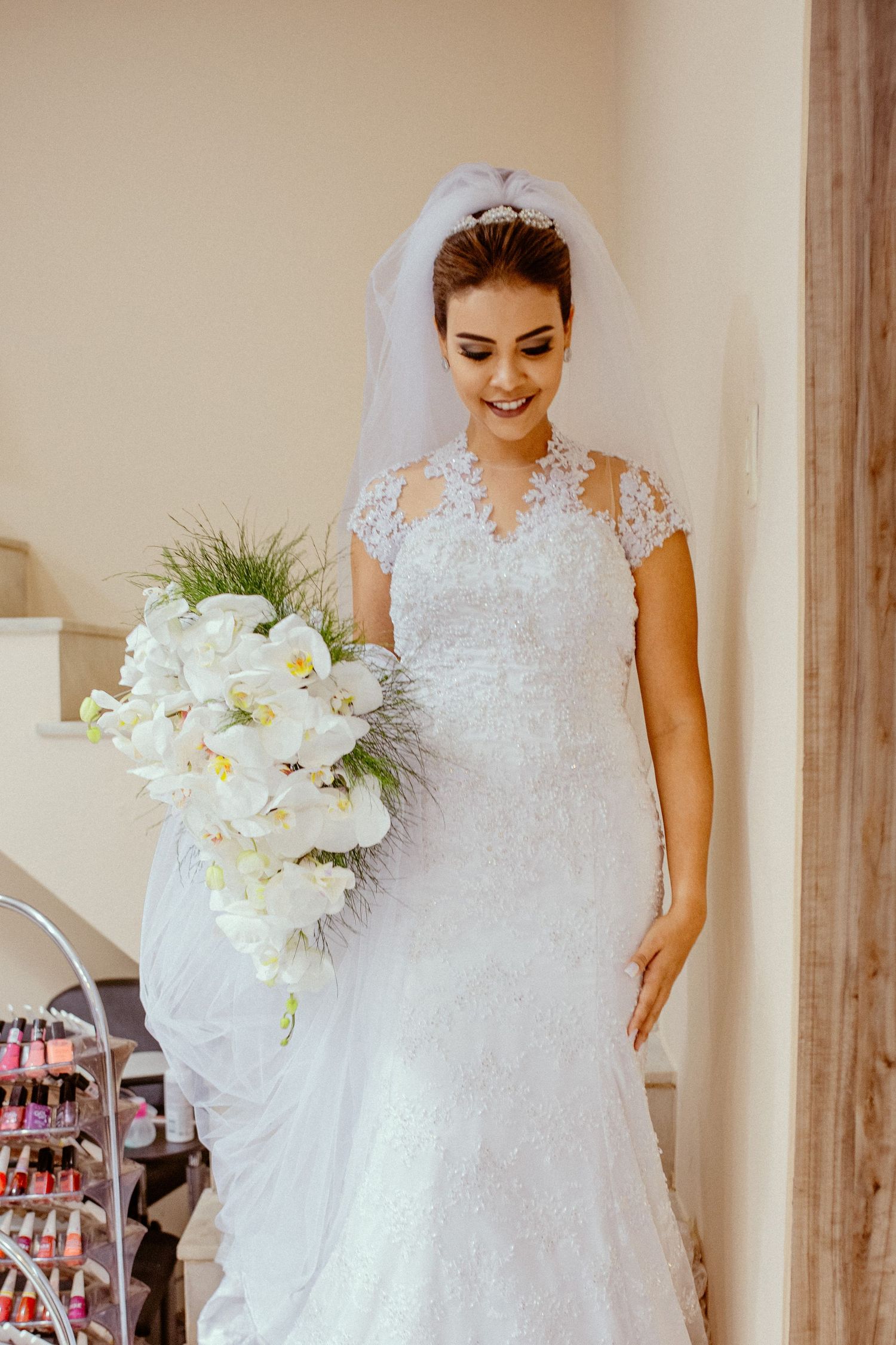 A bride in a lace wedding gown with cap sleeves holds a white orchid bouquet while smiling.