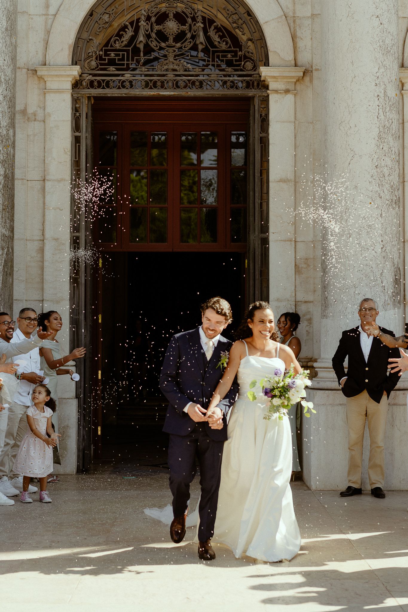 Newlyweds exit church doors as guests shower them with confetti in sunny outdoor celebration.