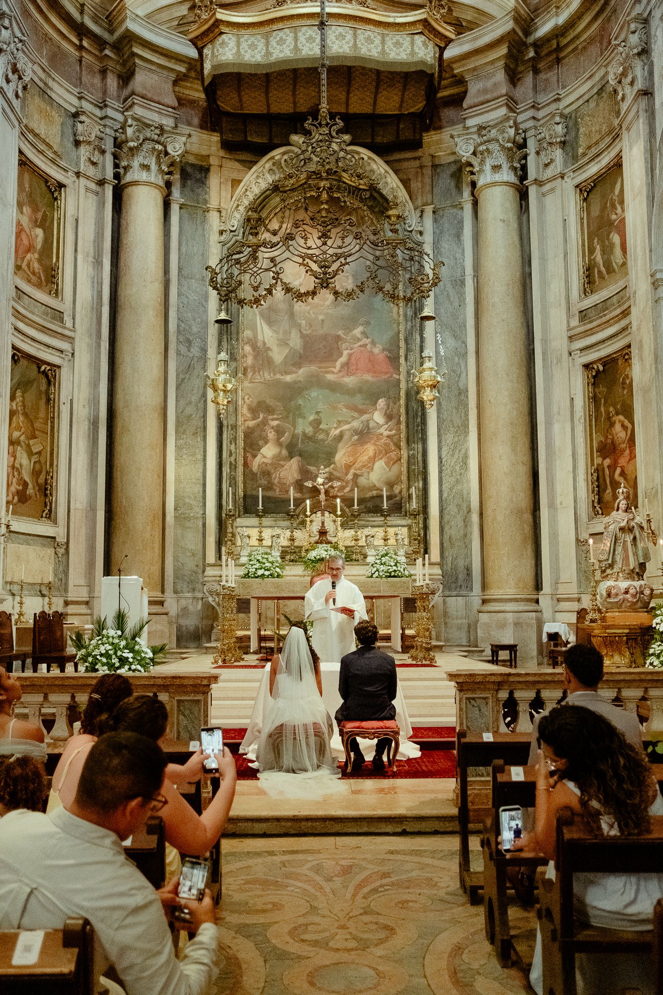 A bride and groom kneel before a priest at a baroque church altar during a traditional Catholic wedding ceremony.