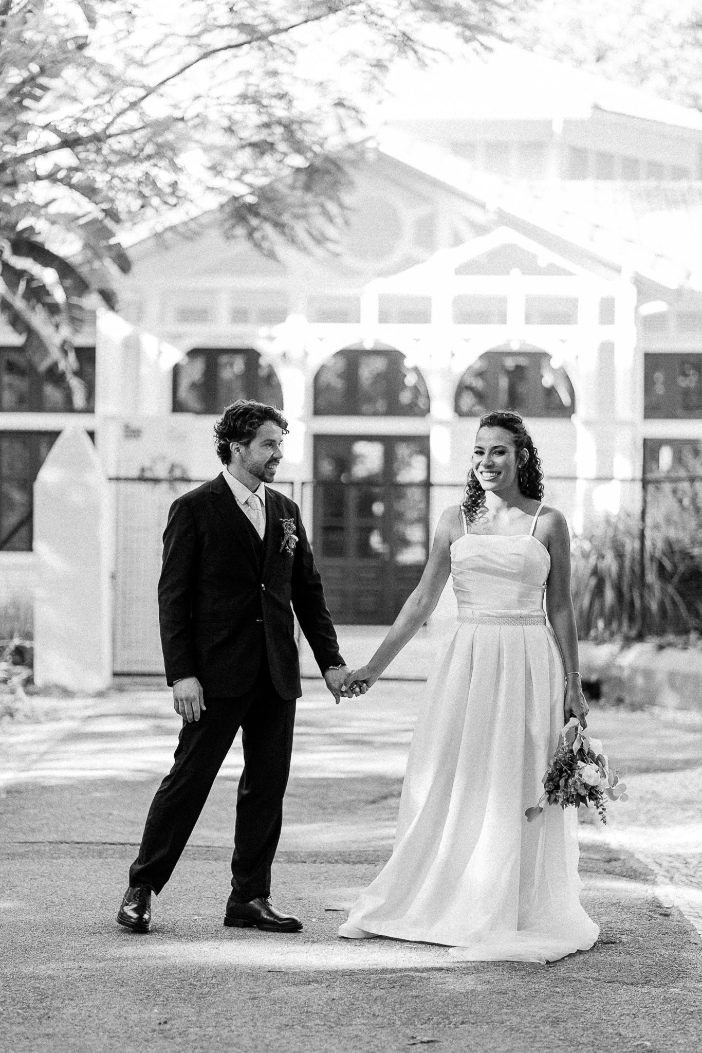 Black and white photo of a bride and groom holding hands, smiling outside a white building on their wedding day.