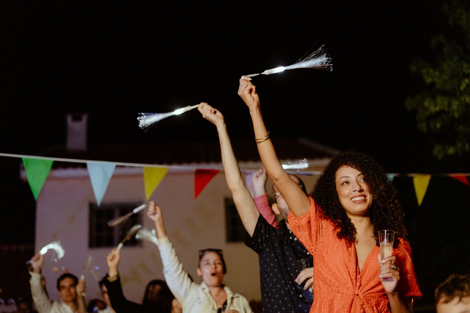 People celebrating outdoors at night, waving light-up sparklers with colorful bunting flags in the background.