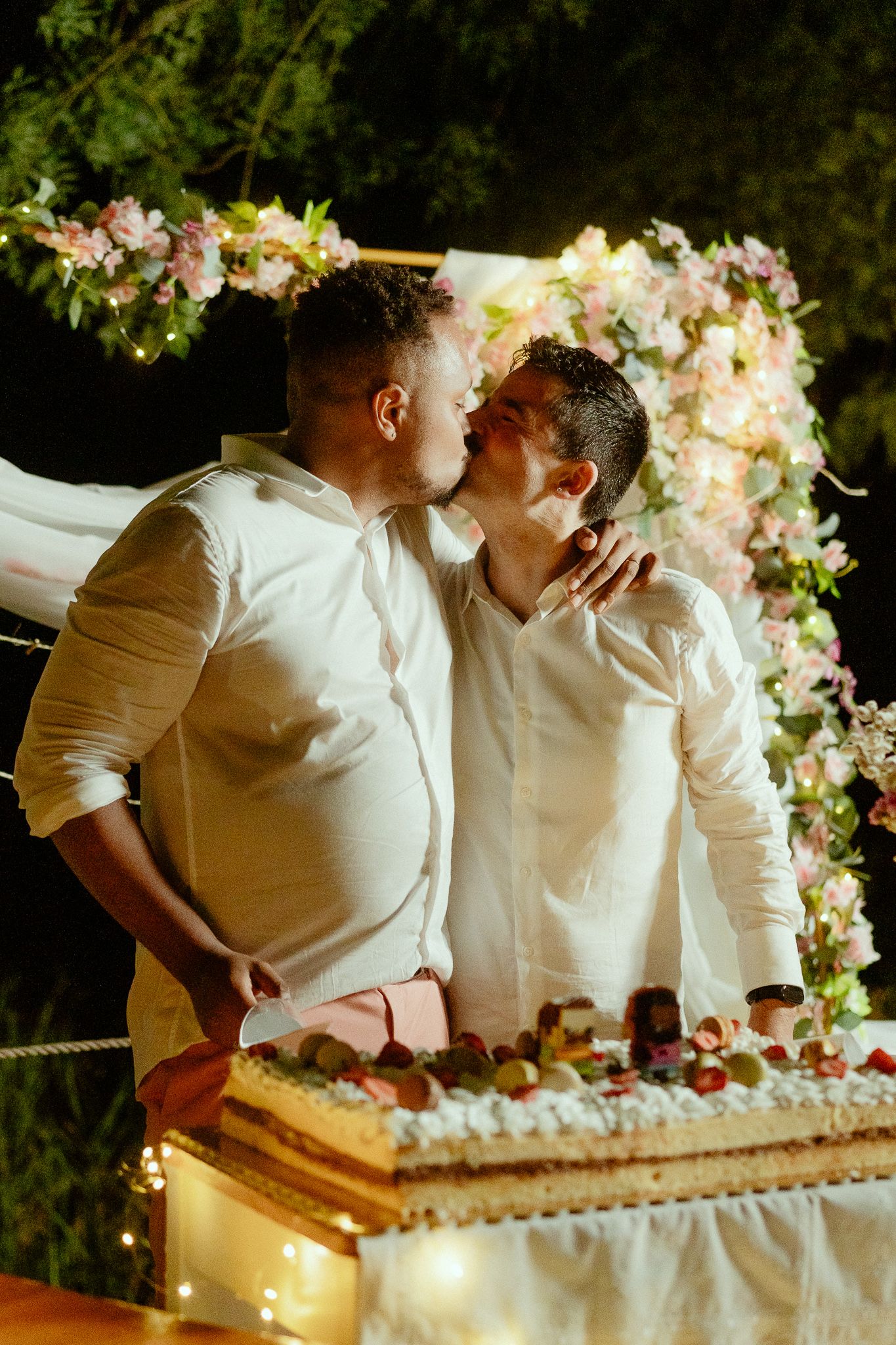 Two grooms sharing a kiss at their wedding reception beside a decorated cake under floral arch.