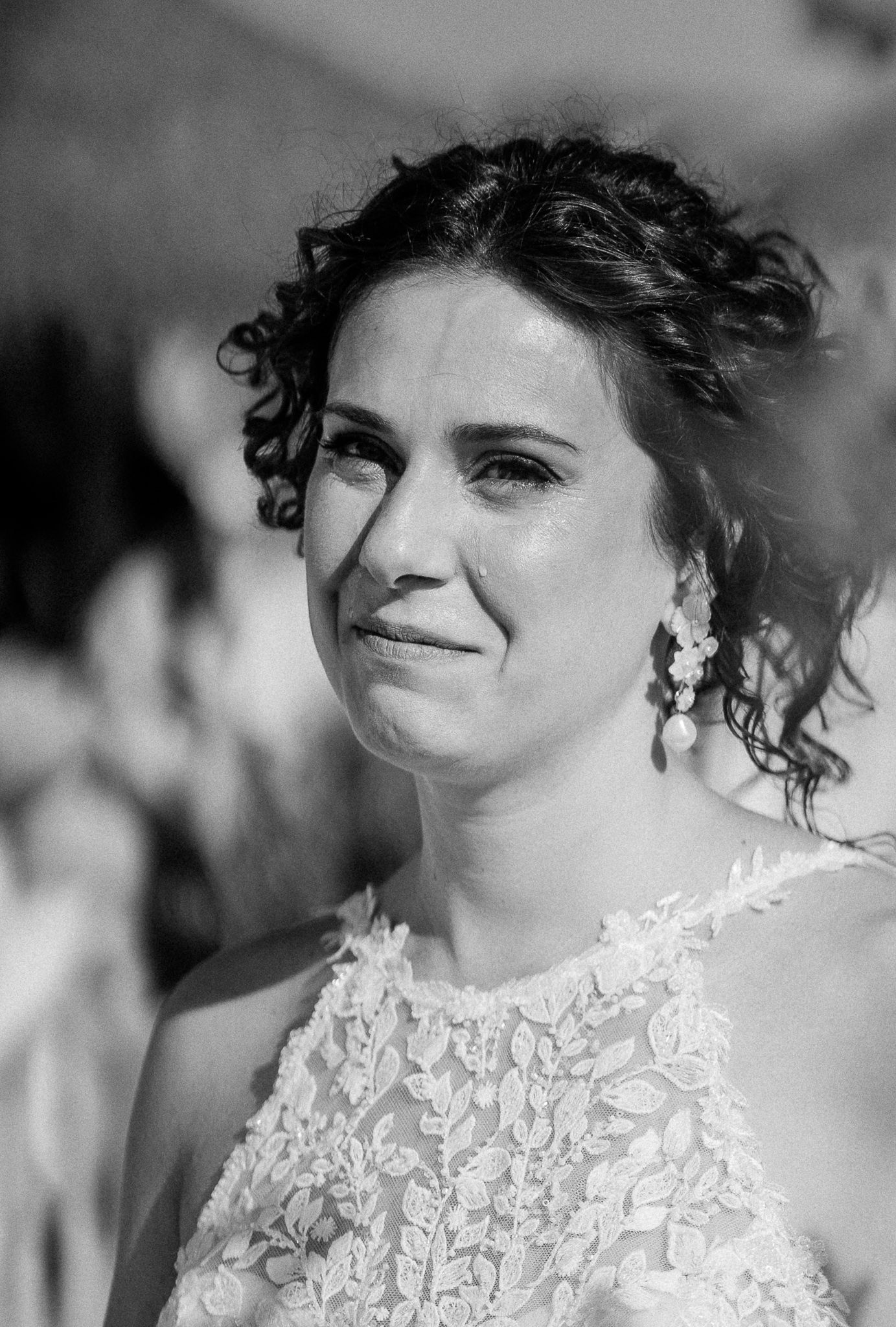 Black and white portrait of a bride with curly updo, pearl earrings, and lace dress, showing emotional expression.