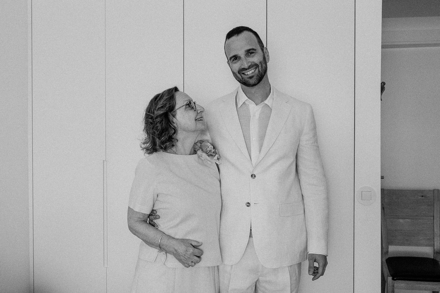 Black and white photo of a smiling couple in elegant attire standing together against a white paneled wall.