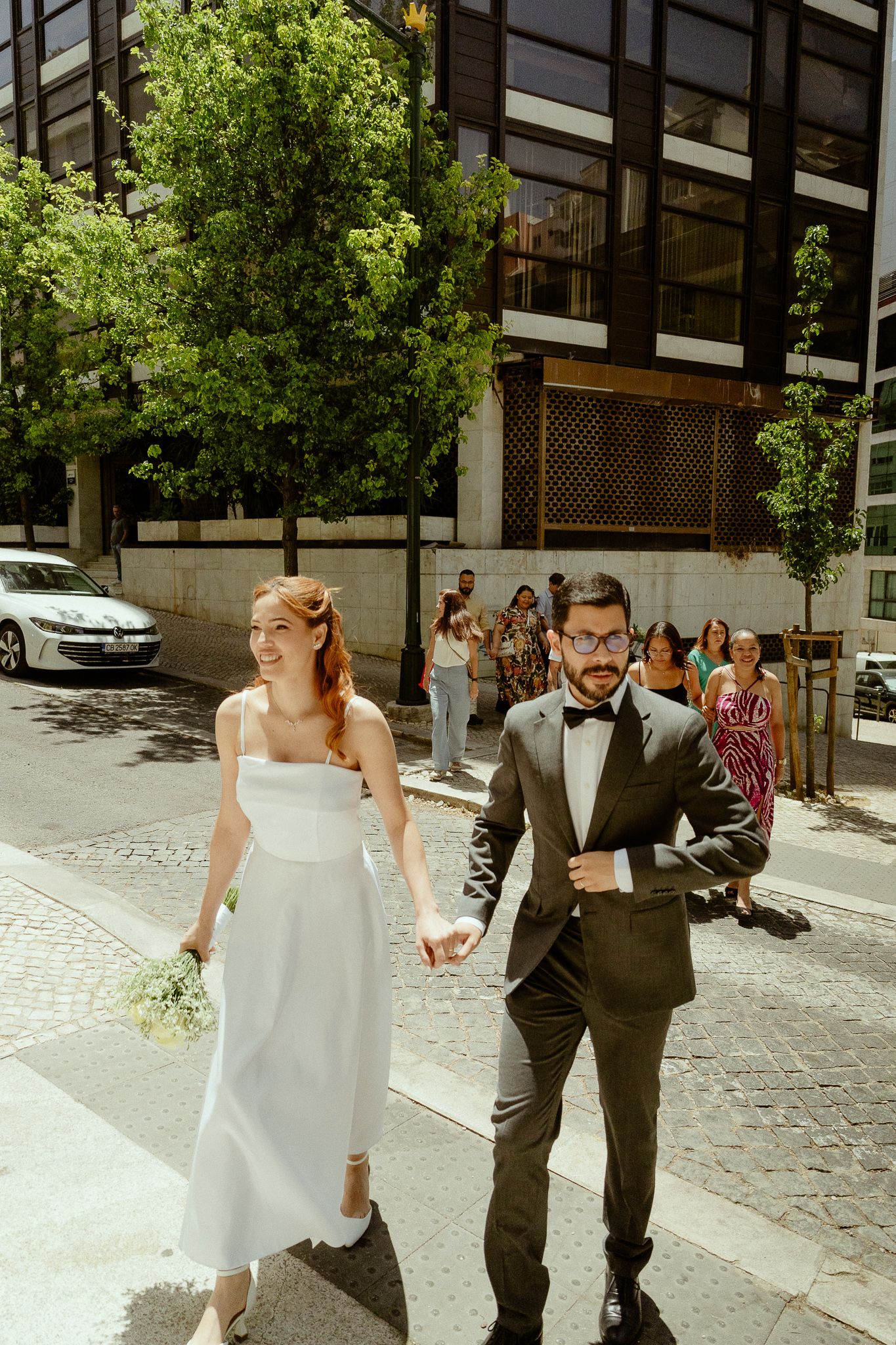 Bride in white dress and groom in dark suit hold hands walking down a sunny city street after their wedding ceremony.