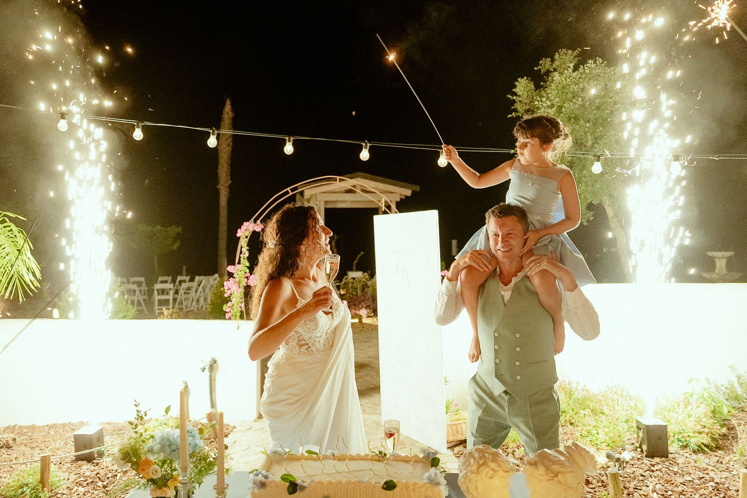 Family celebrating at wedding with sparklers, cake, and fireworks at night outdoor venue.