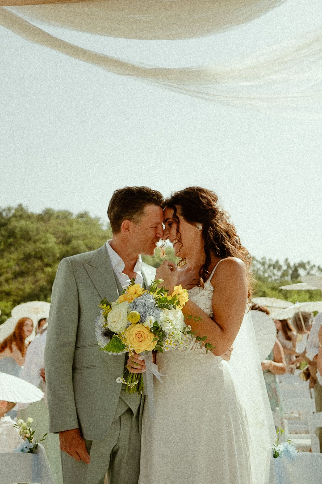 Bride and groom touching foreheads at outdoor wedding ceremony, bride holding yellow and white bouquet, groom in gray suit.