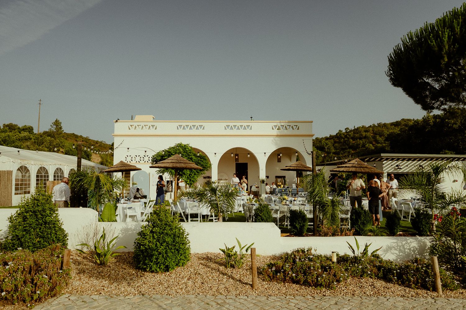 Elegant white Mediterranean villa wedding venue with outdoor dining tables, lush greenery, and guests gathered under clear blue skies.