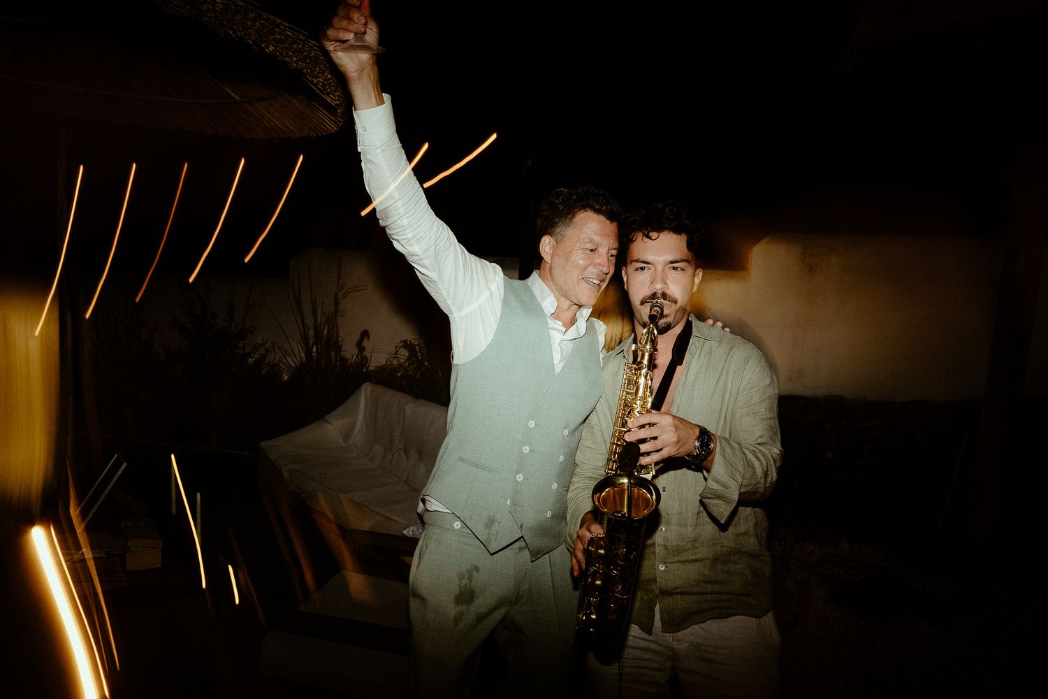 Two men in light suits celebrate at night, one raising his arm while the other plays a saxophone.