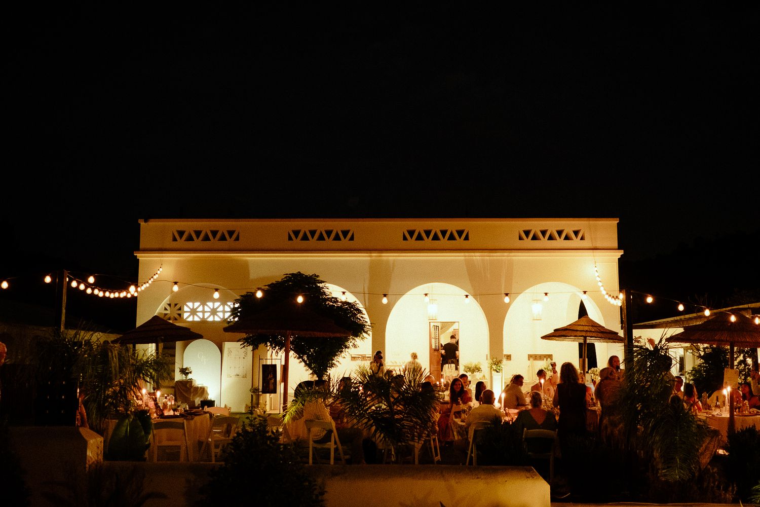 Elegant outdoor wedding reception at night with string lights illuminating a white arched venue.