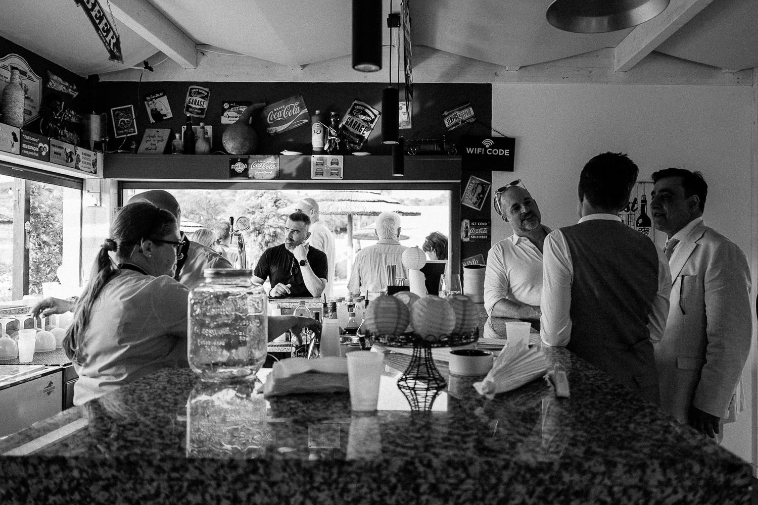 Black and white photo of people gathered at a bar counter inside a casual venue with snack displays in the background.