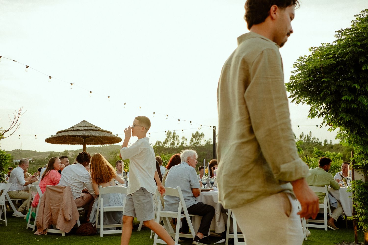 Outdoor garden party with guests dining at white chairs under string lights at dusk.