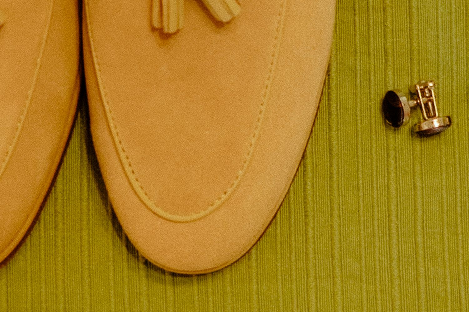Tan suede loafer shoes with tassel detail beside gold cufflinks on a green striped background.