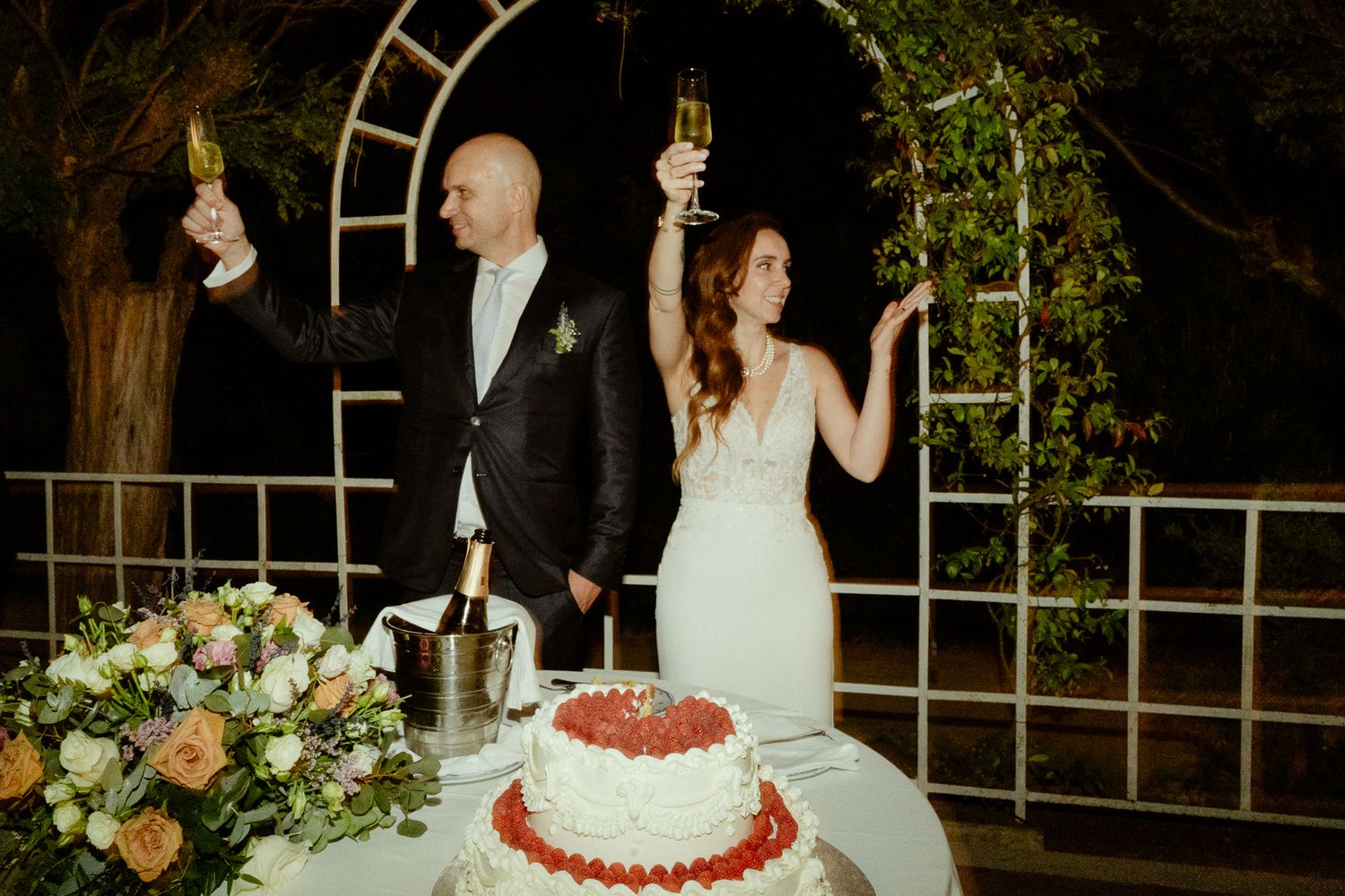 Bride and groom toasting with champagne beside a wedding cake adorned with strawberries and flowers.