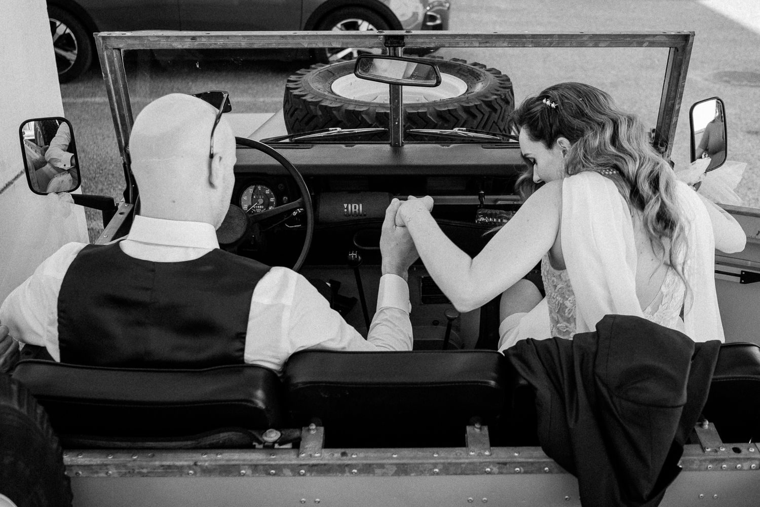 Black and white photo of a couple holding hands in a vintage open-top vehicle, dressed in wedding attire.