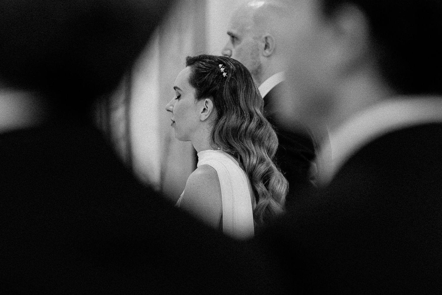 Black and white wedding photo of bride with elegant updo hairstyle, standing between guests during ceremony.